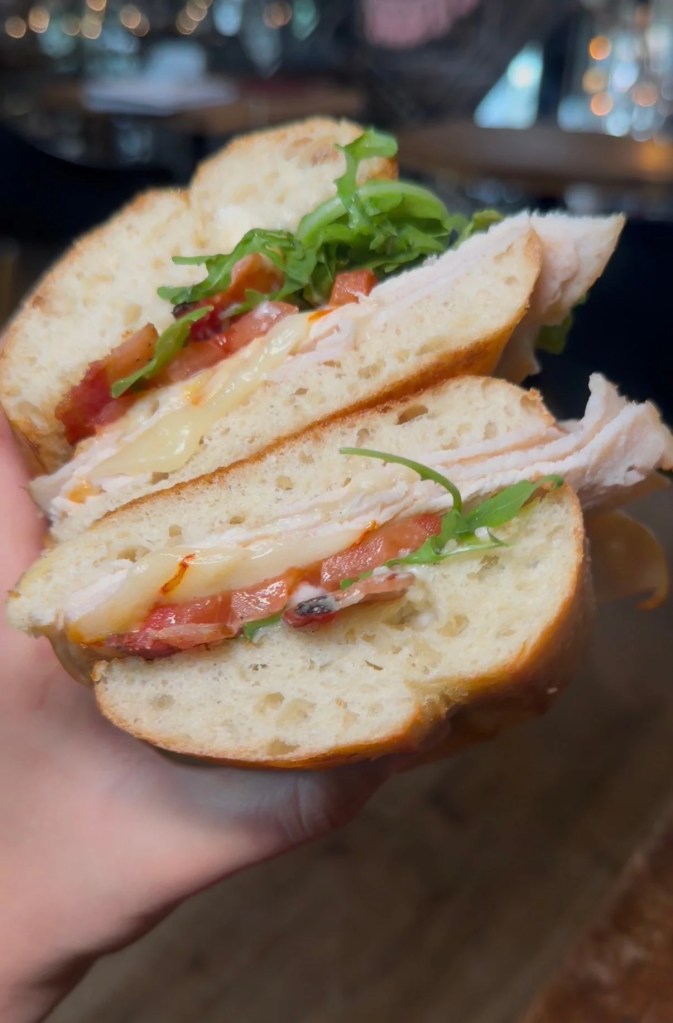 A hand holding two sandwich halves with sliced deli meat, cheese, tomato and leafy greens at Better Buzz.