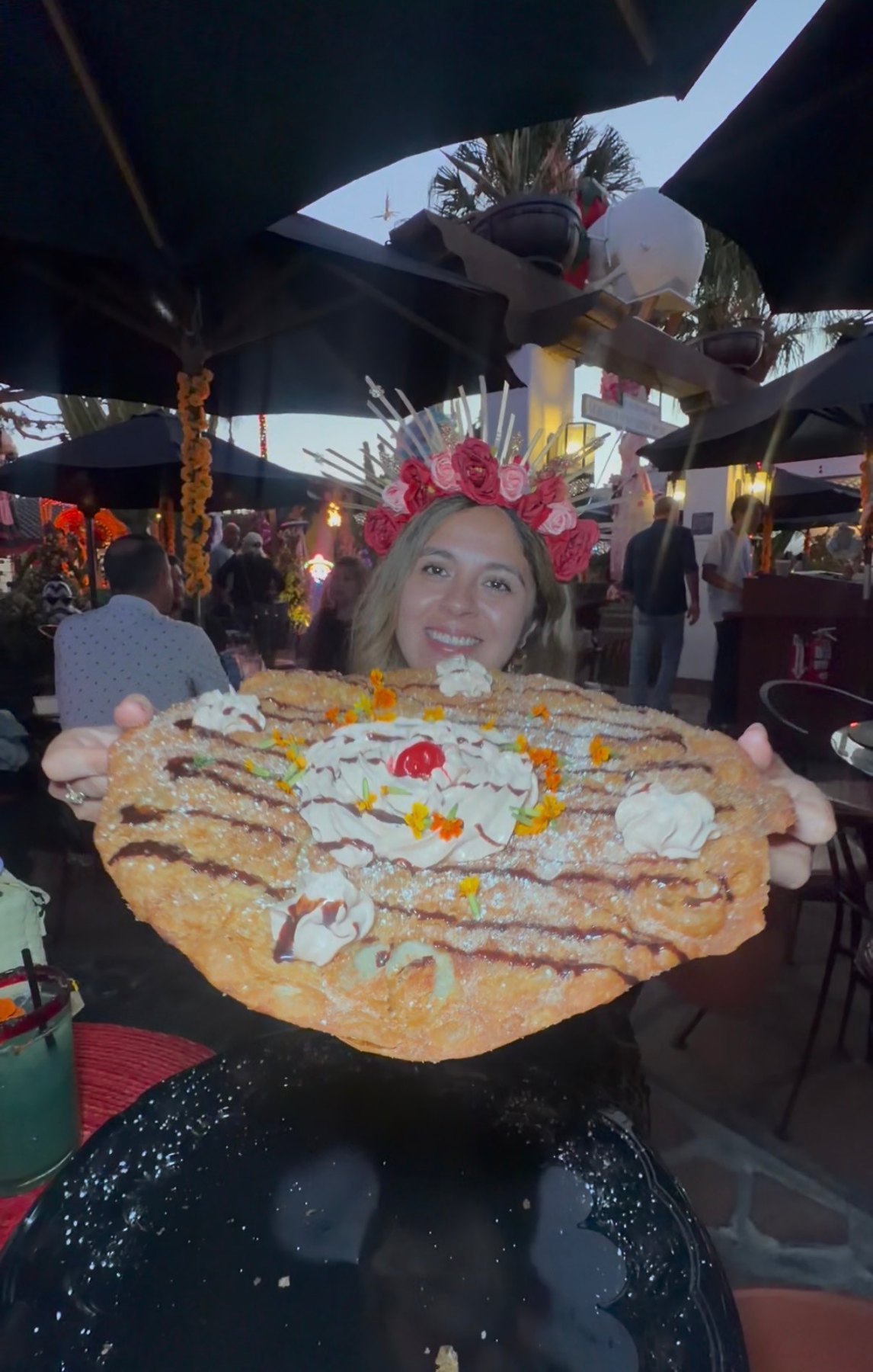 A woman in a pink flower crown holds a giant buñuelo topped with cream, a cherry and colorful petals at Casa de Reyes.