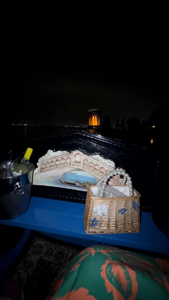 A glowing lantern, a Venice bridge decoration, a bottle in an ice bucket and a woven handbag aboard a gondola at night.