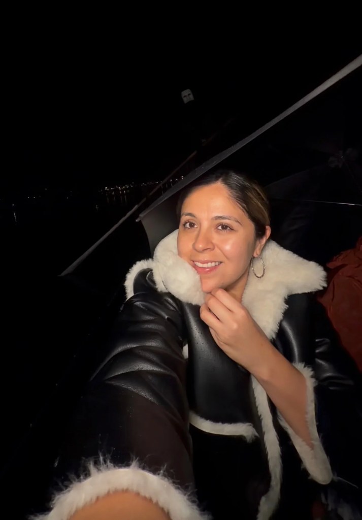 A woman in a black jacket with cream trim smiles aboard a gondola at night, with a masked gondolier behind her.