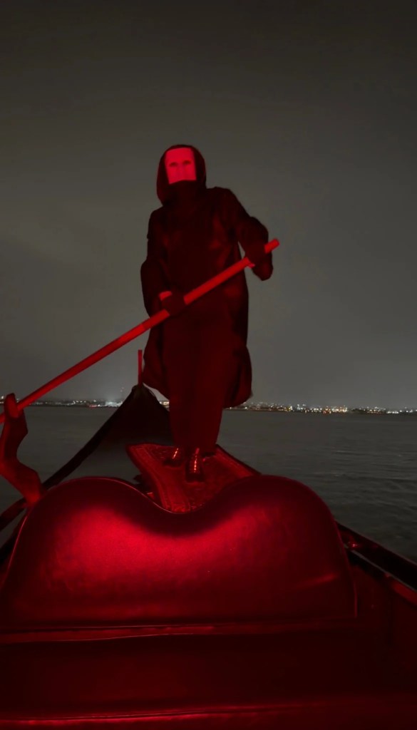 A masked phantom gondolier rows a red-lit gondola on the water at night in Coronado.