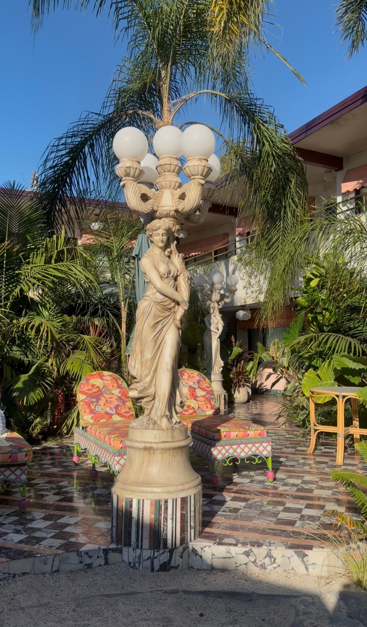 A sculpted lamp post, colorful chairs and palm trees in a tiled courtyard.