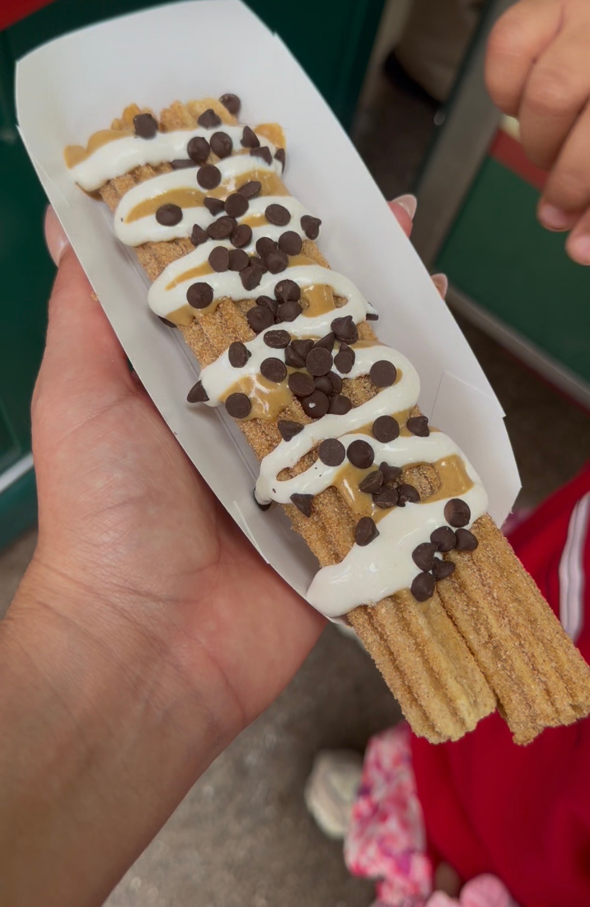 A churro in a paper tray topped with white and tan drizzle and chocolate chips.