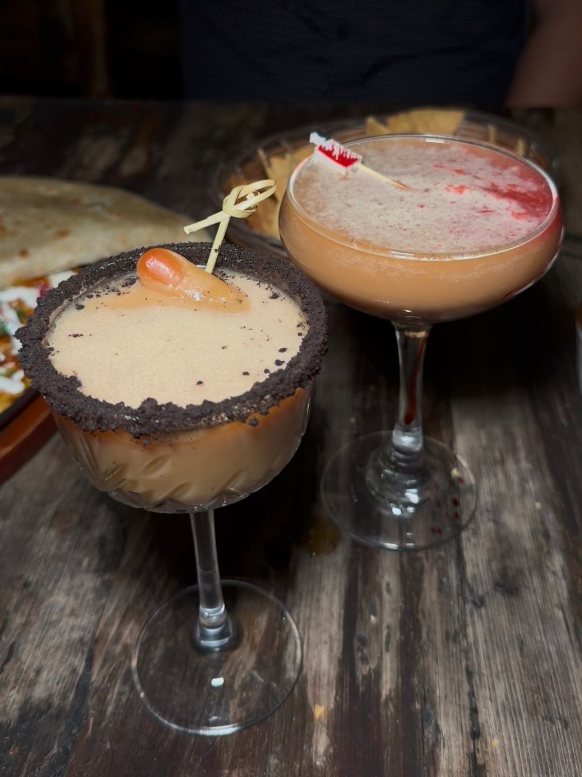 Two Halloween-themed cocktails in stemmed glasses on a wooden table at El Sueño.