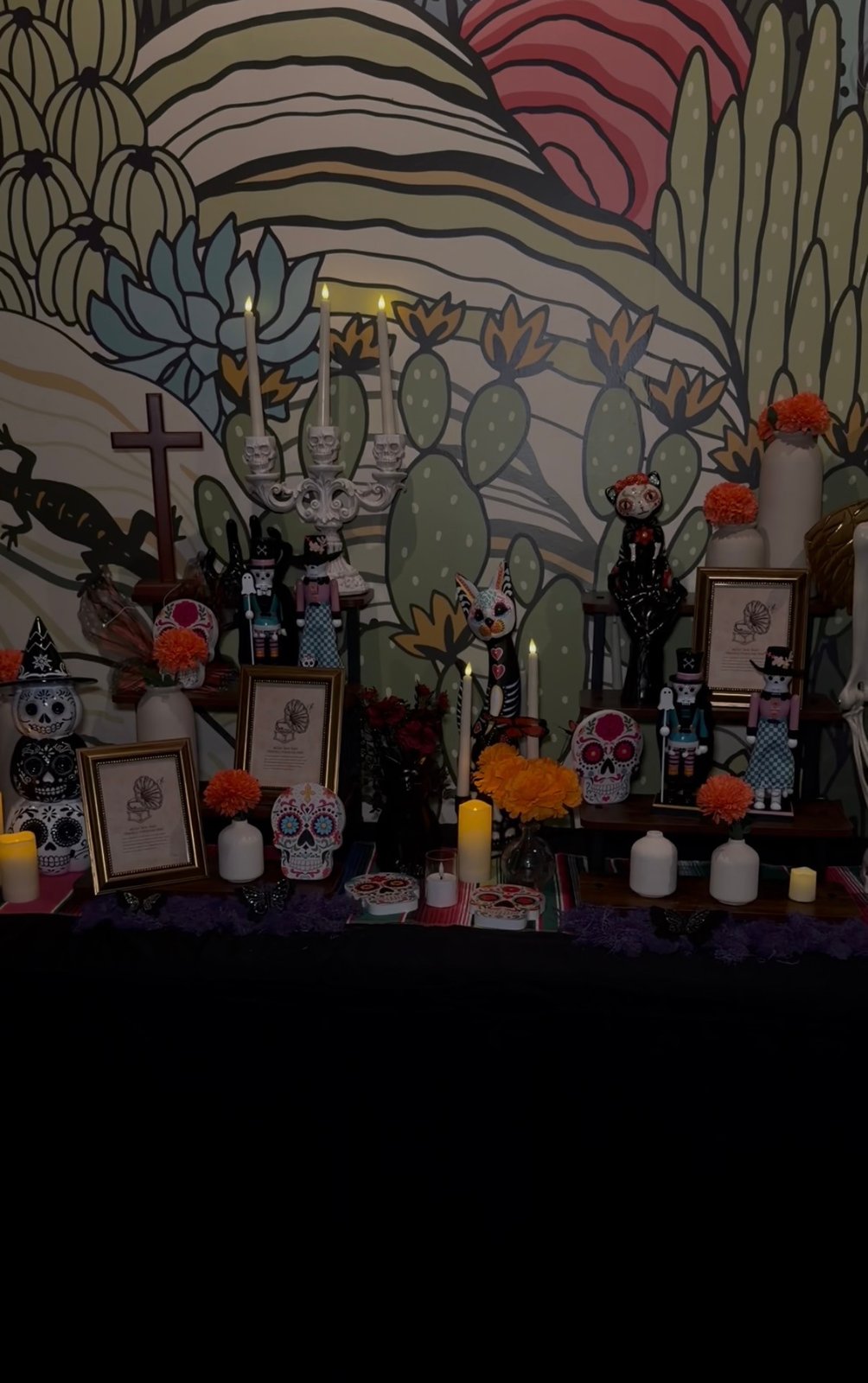 Día de los Muertos display with painted skulls, skeleton figures, marigolds and candles in front of a cactus mural at El Sueño.