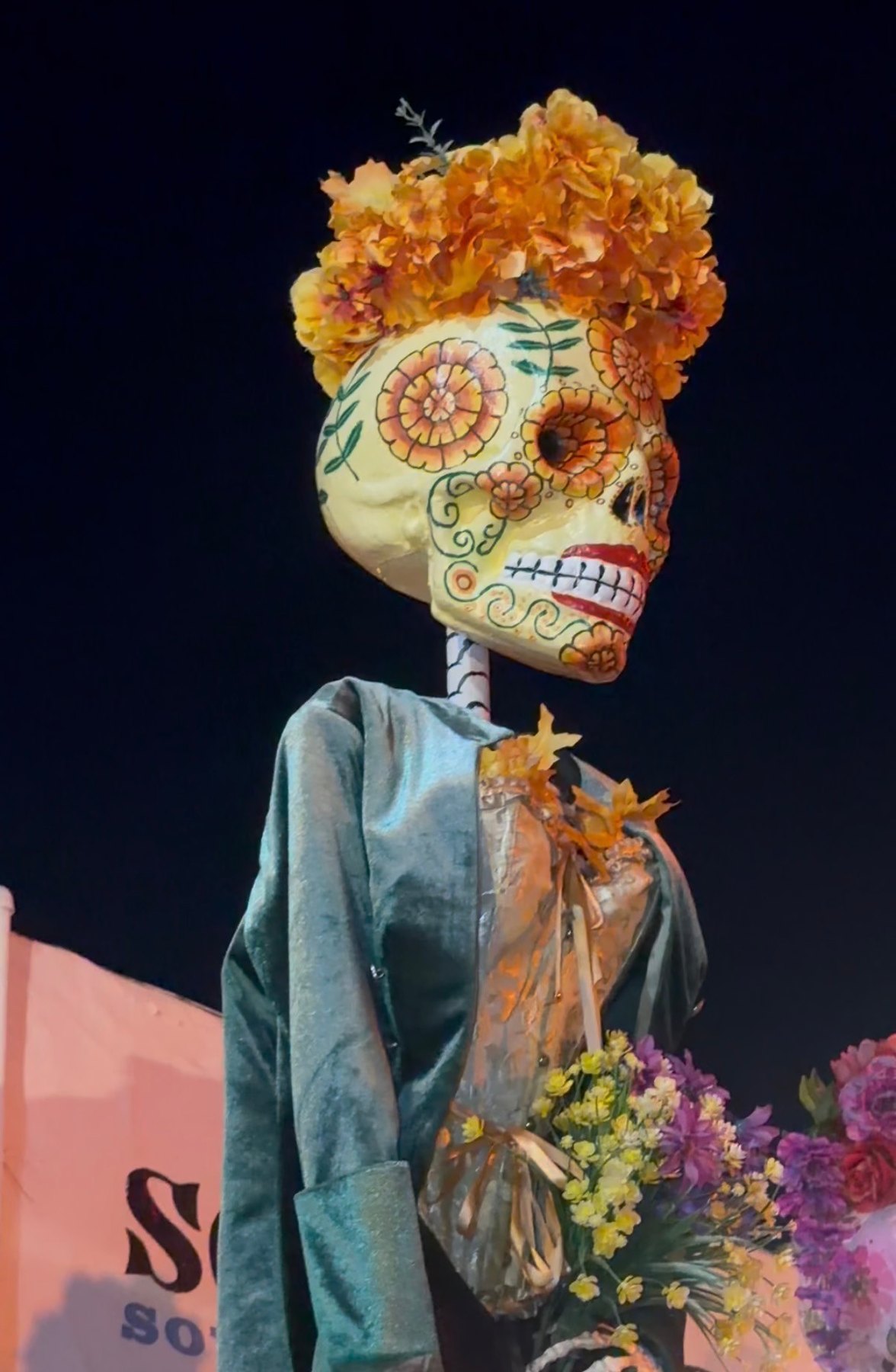 A Catrina with a painted floral skull, orange flower crown and green shawl in an evening Día de los Muertos display.