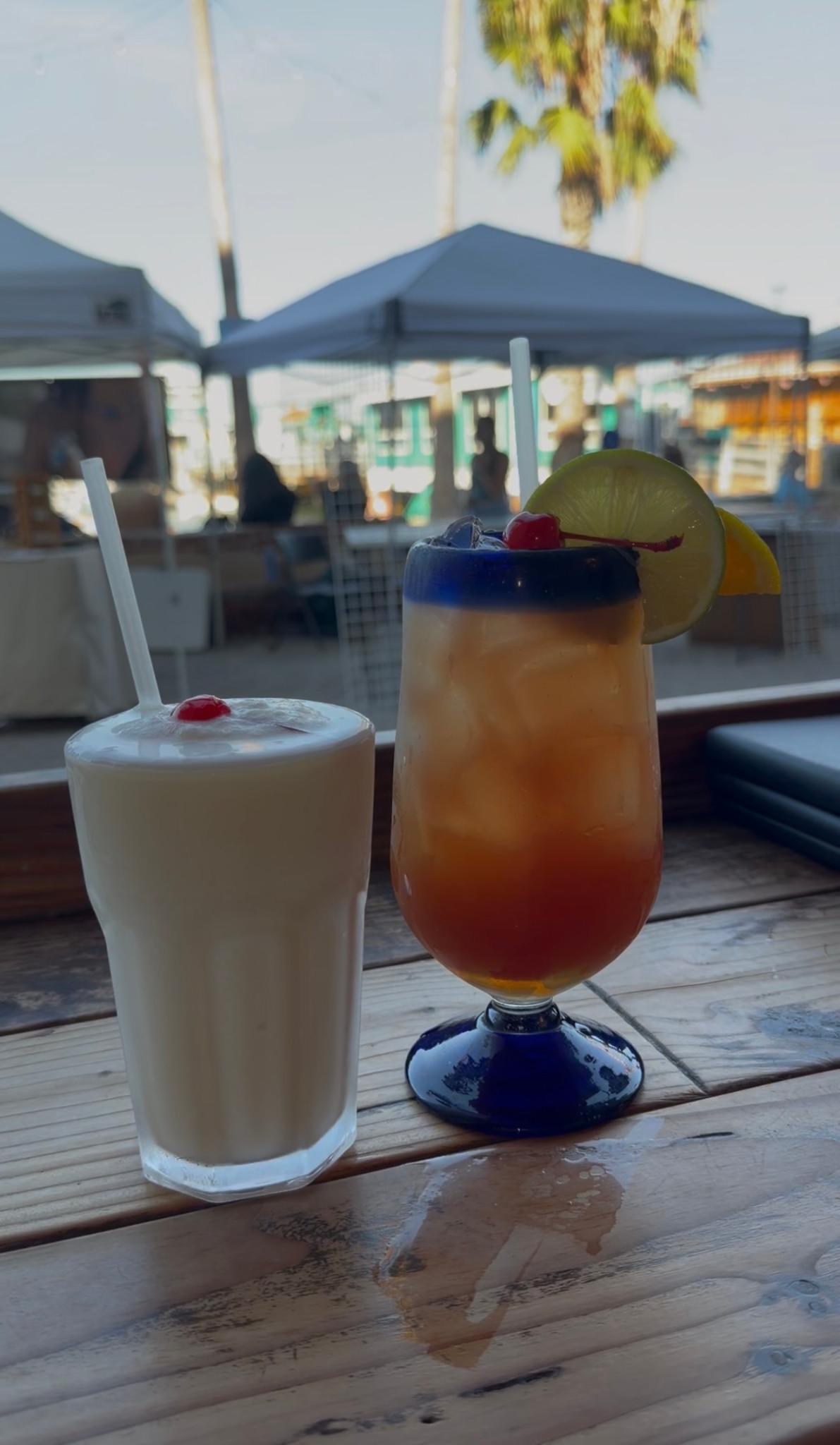 Two colorful cocktails on a wooden table with patio umbrellas and palm trees outside.