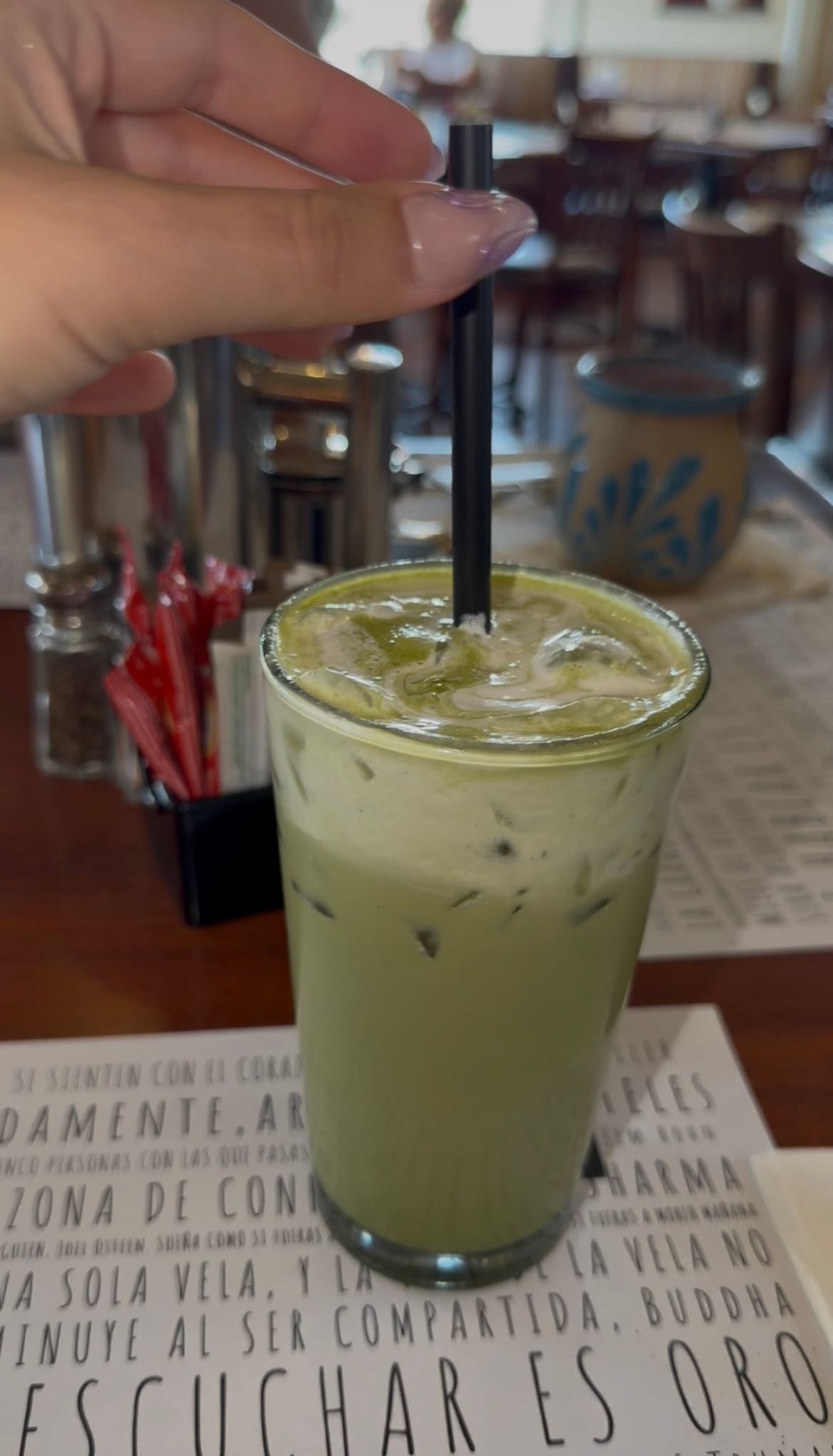 An iced pale-green drink in a tall glass with a black straw on a Café de la Flor table.