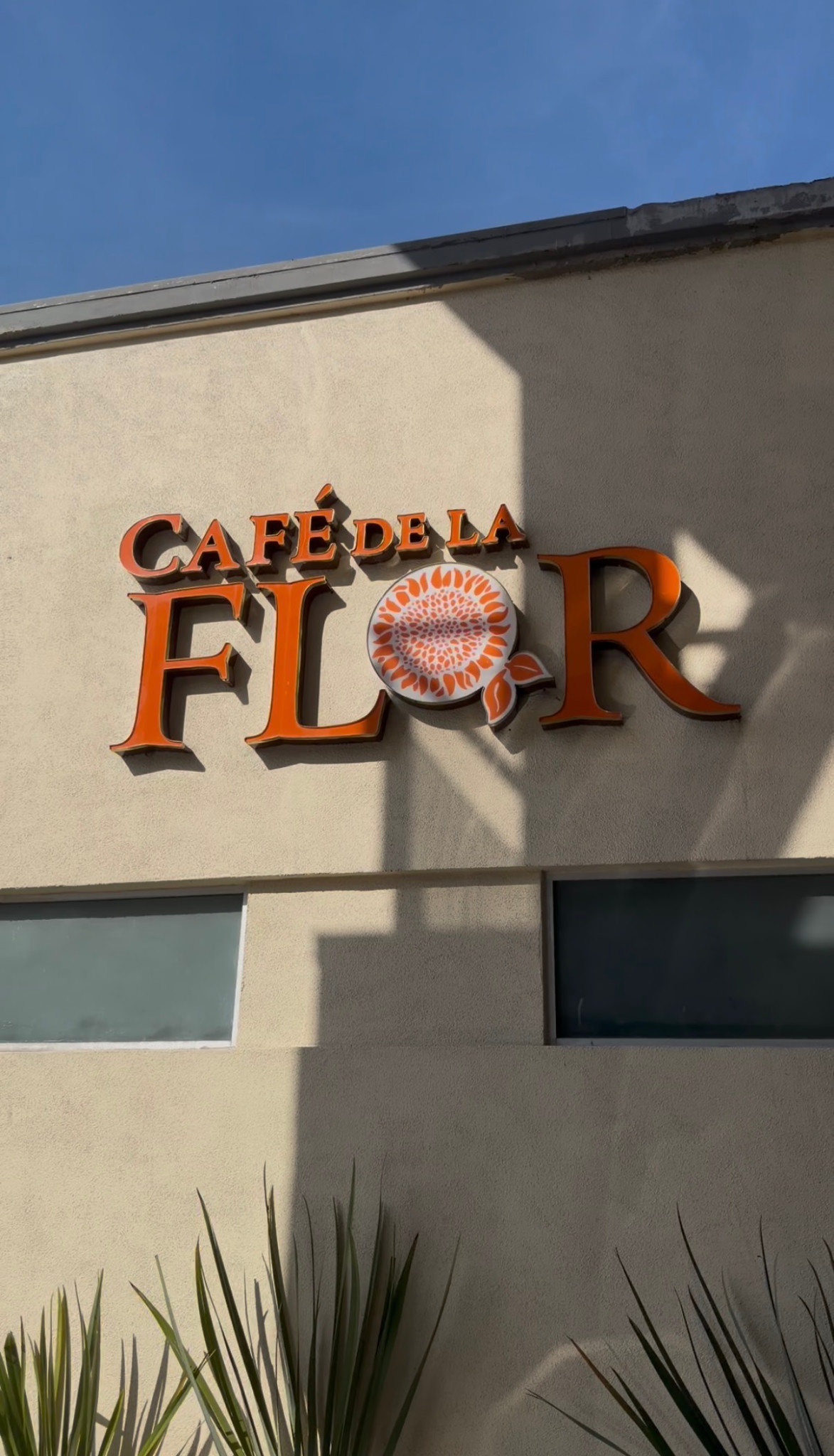 Orange Café de la Flor lettering and a floral emblem on a cream-colored exterior wall.