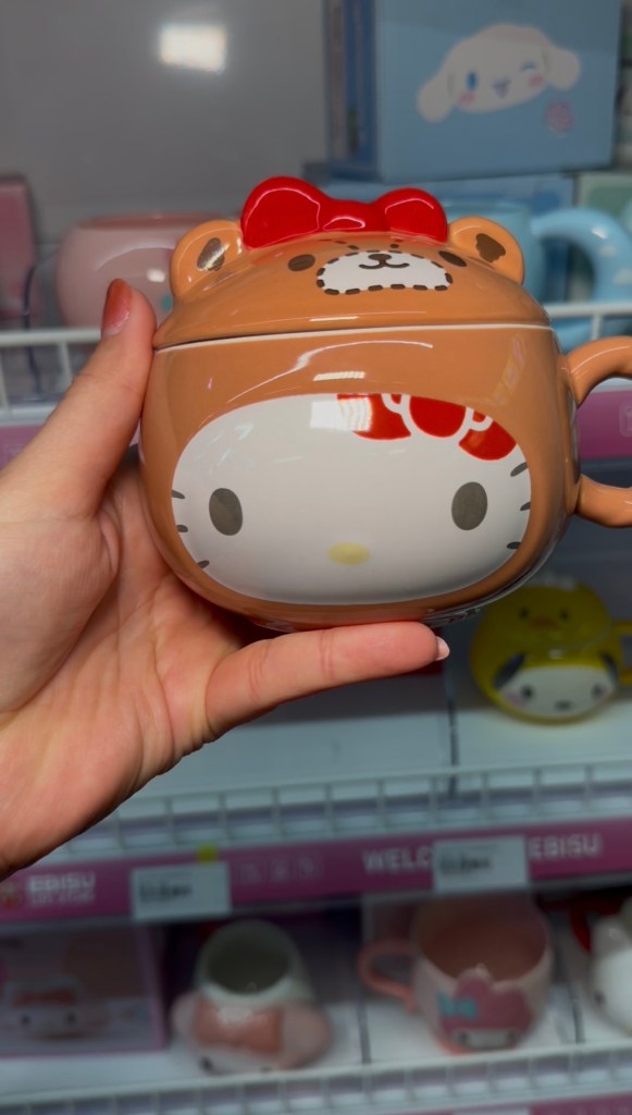 A hand holds a brown Hello Kitty mug with a matching lid and red bow in front of character mugs at EBISU.