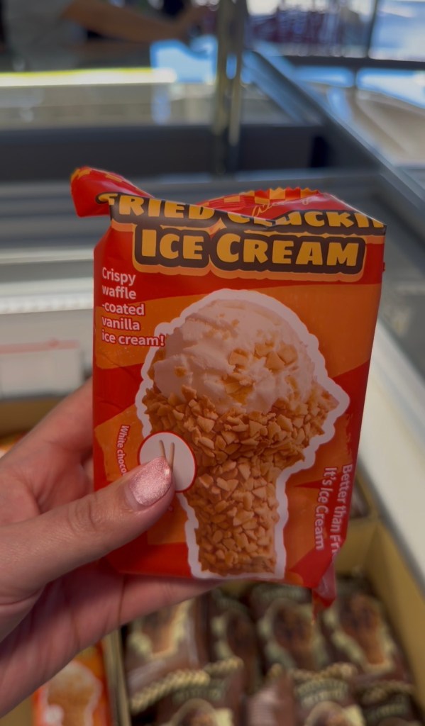 A hand holds an orange package labeled Fried Chicken Ice Cream above a store freezer.