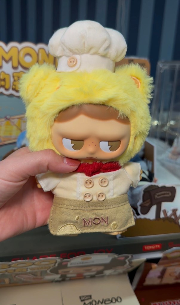 A hand holds a yellow plush collectible wearing a white chef hat, jacket and tan apron at EBISU.