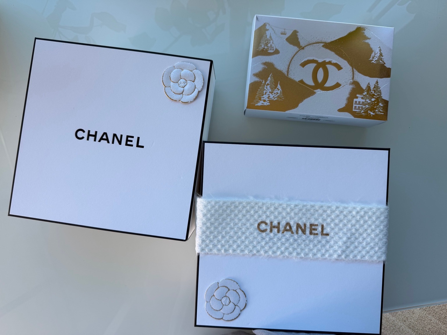 Chanel white gift boxes with camellia details, a white knit band and gold illustrated packaging on a glass table.