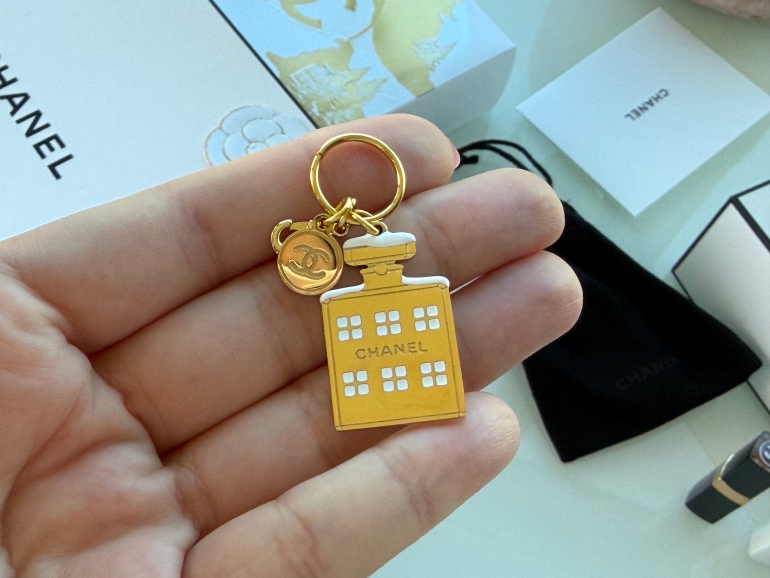 A hand holding a gold-tone Chanel bottle-shaped charm with white window-like details.