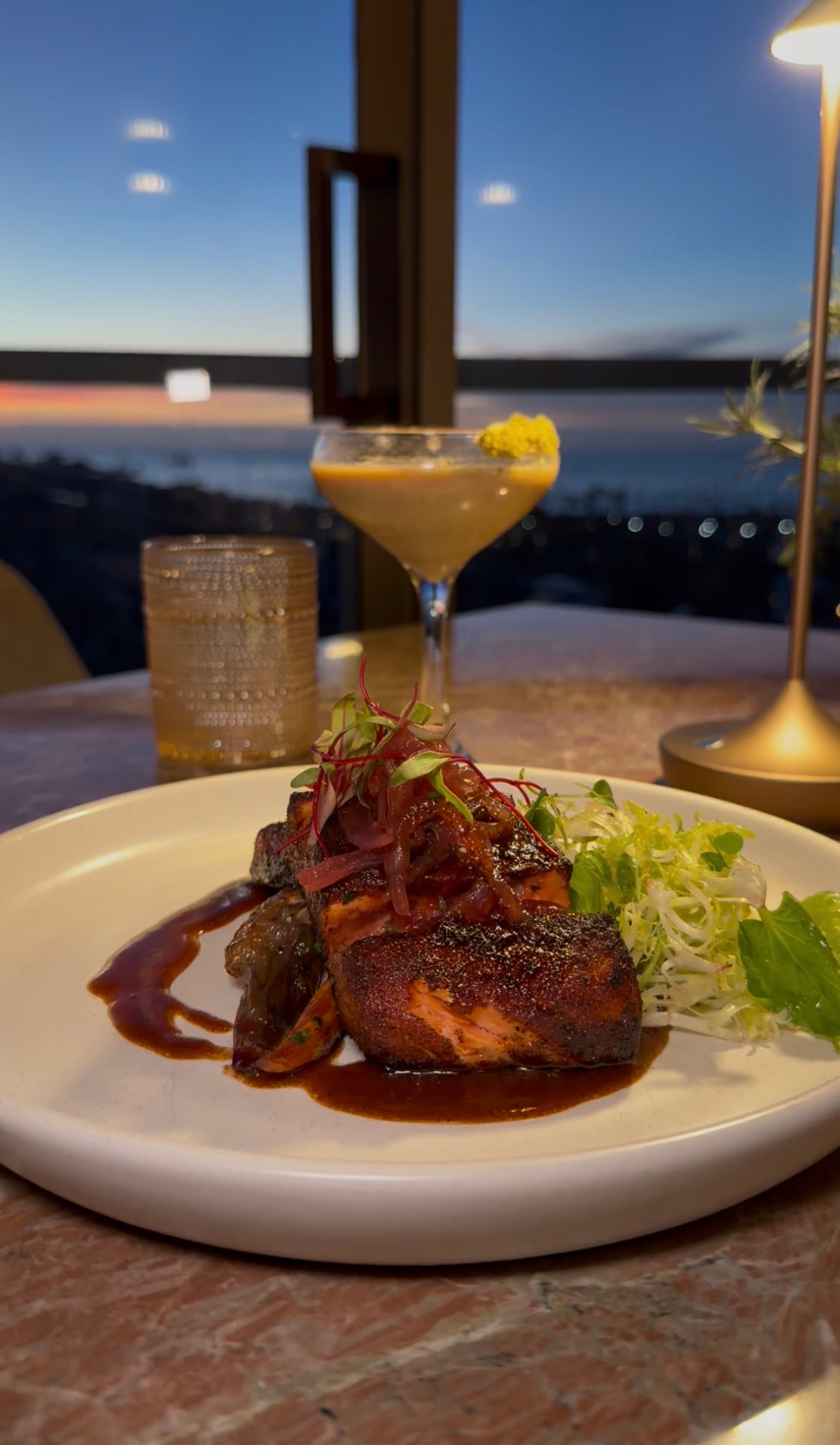 A plated dish and cocktail beside the ocean-view windows at Aries La Jolla.