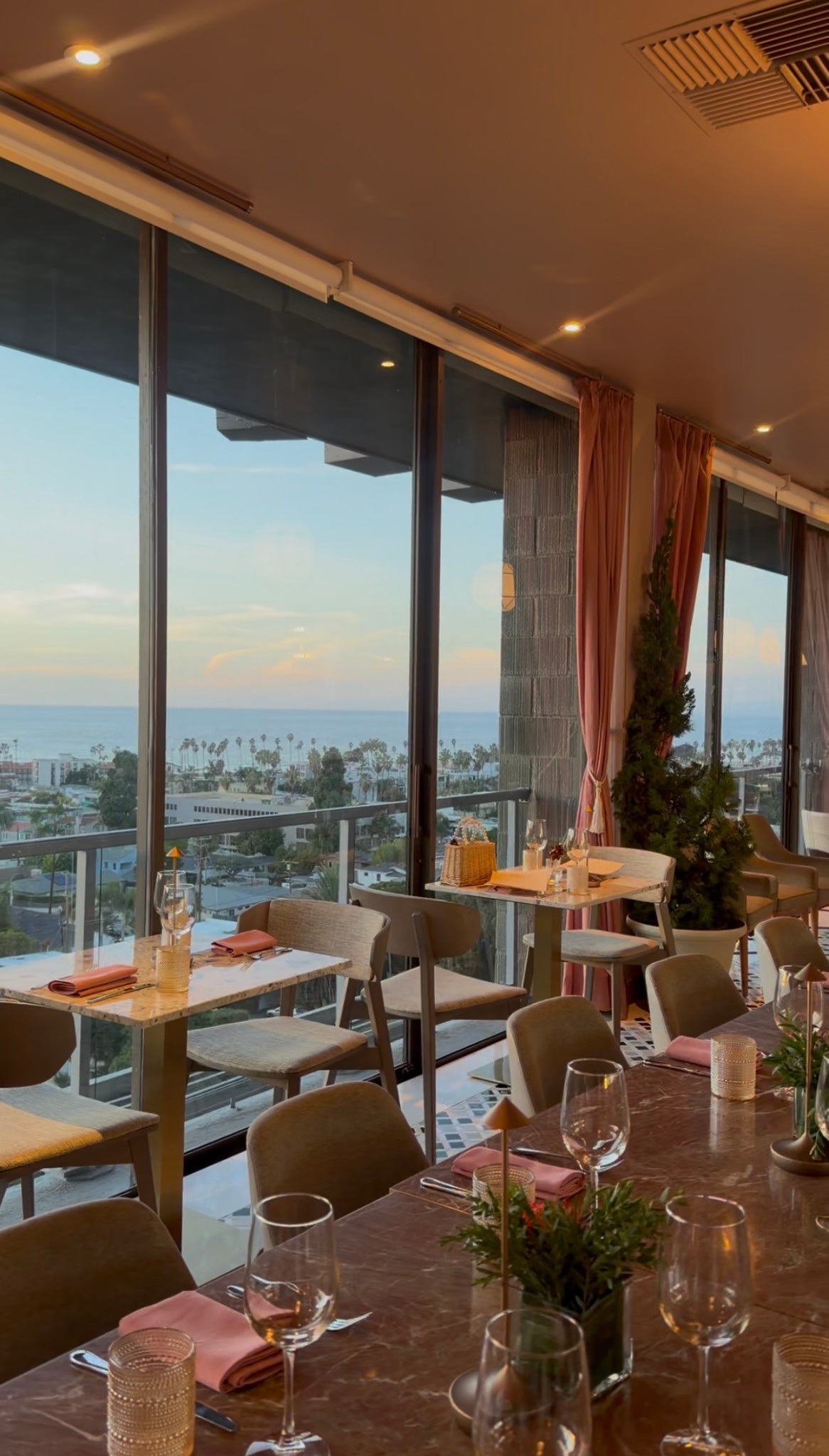Window-side tables and pink curtains overlooking the ocean at Aries La Jolla.