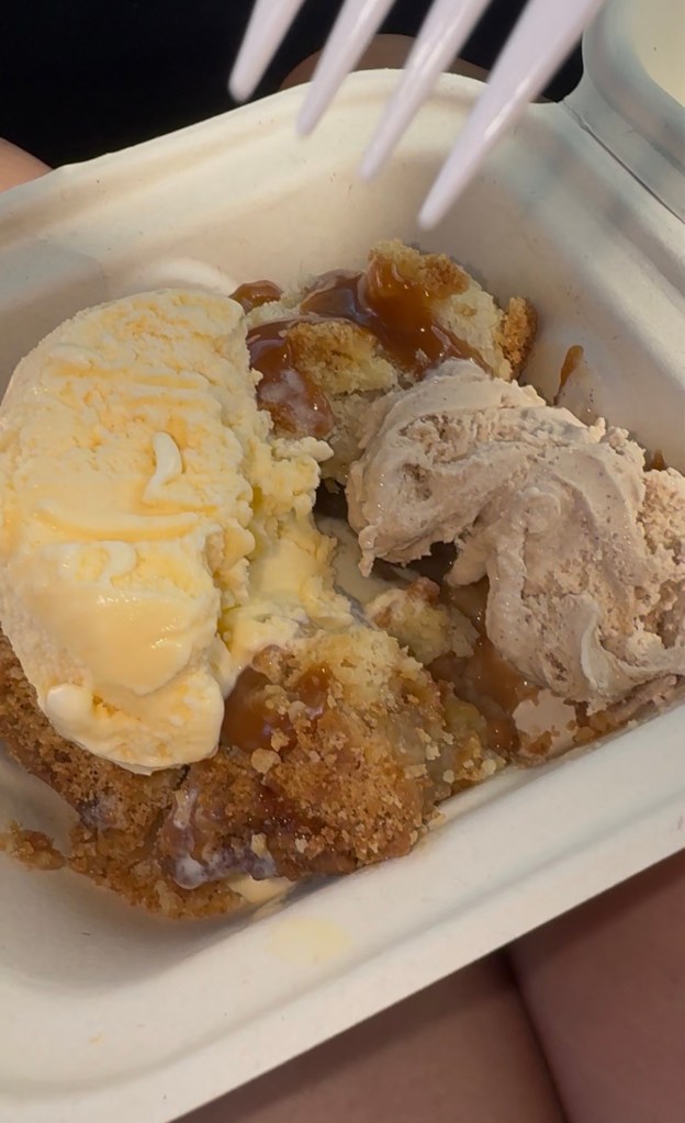 Close-up of crumb-topped pie with two scoops of ice cream and sauce in a takeaway tray.