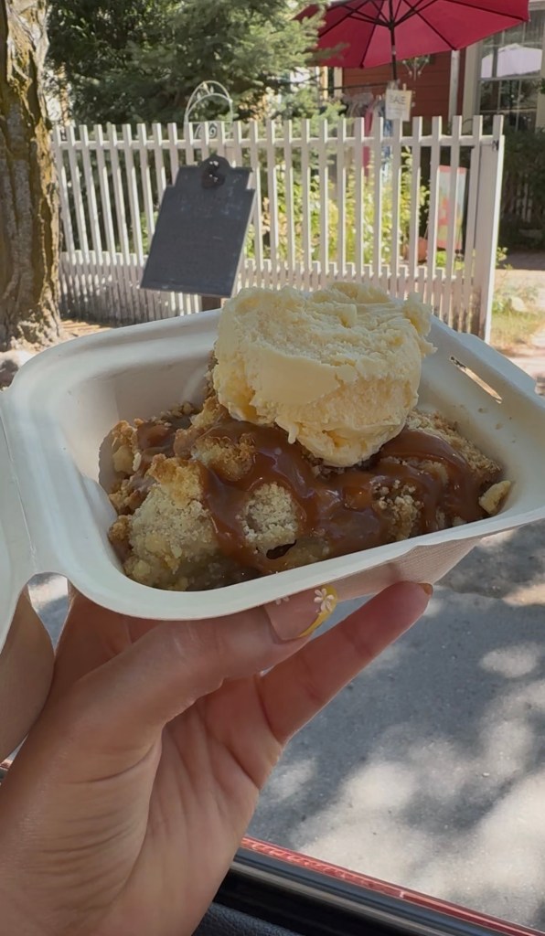 A hand holding a tray of pie topped with ice cream, with a white picket fence and red patio umbrella behind it.