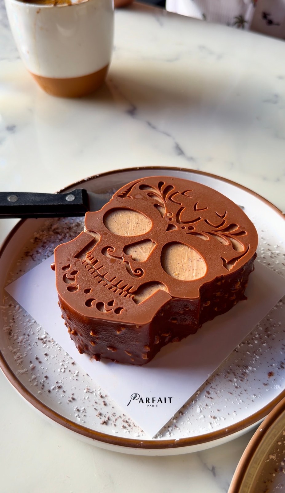 Skull-shaped cake with an intricate brown calavera topper on a Parfait Paris cake board.