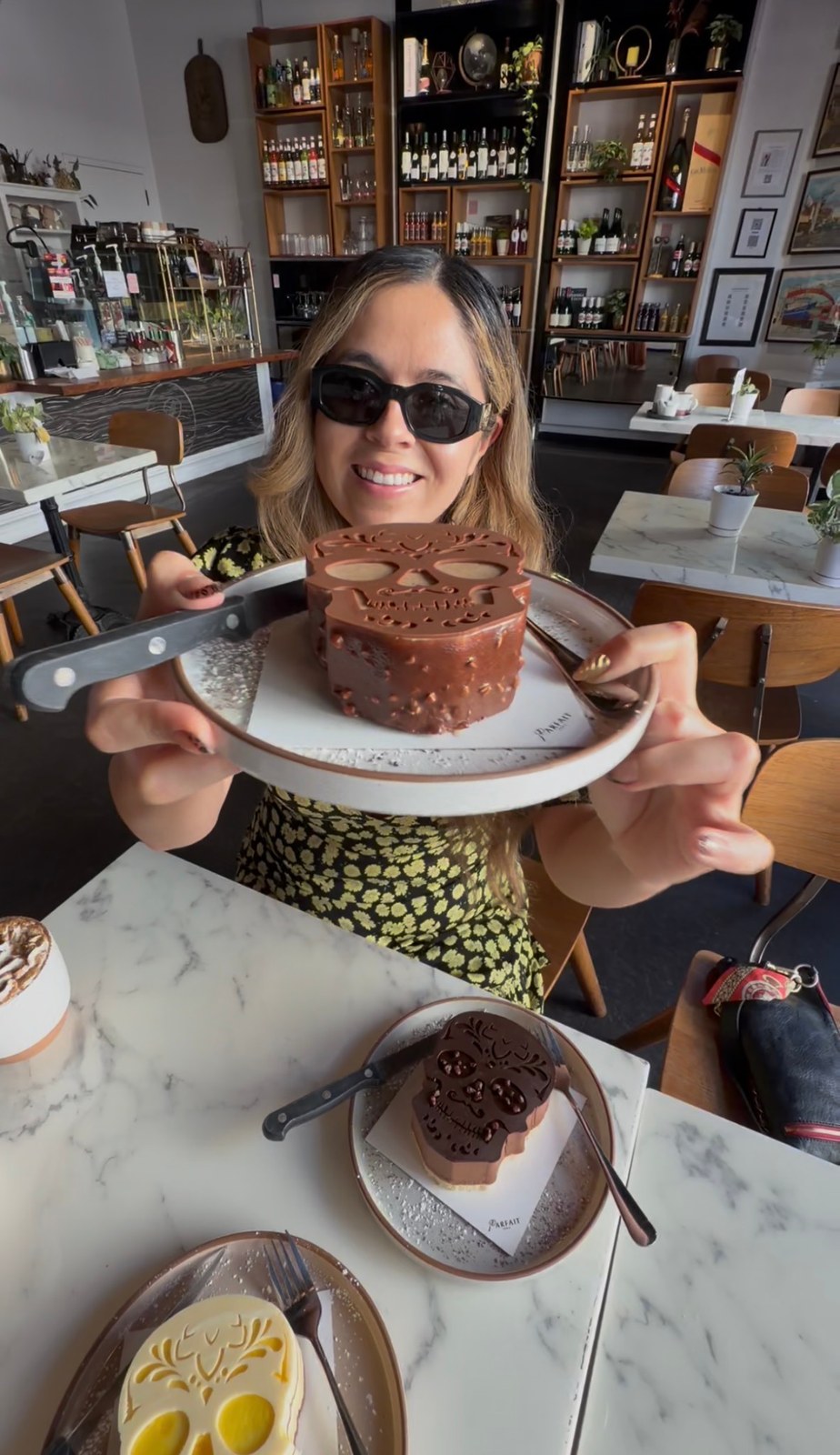 A woman holds a plate with a skull-shaped cake above a table with two more calavera cakes.