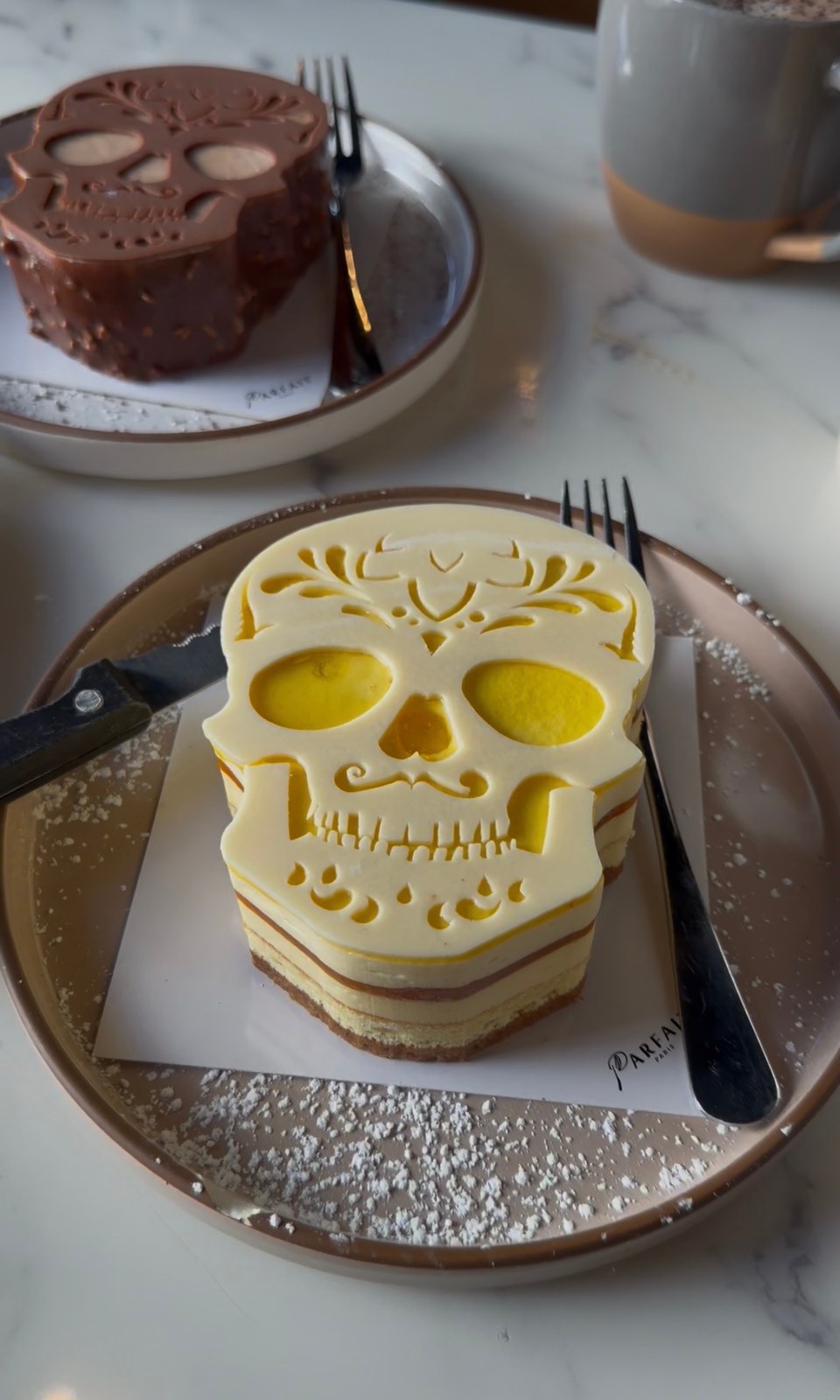 Layered skull-shaped cake with a cream-colored calavera topper and yellow accents at Le Parfait Paris.