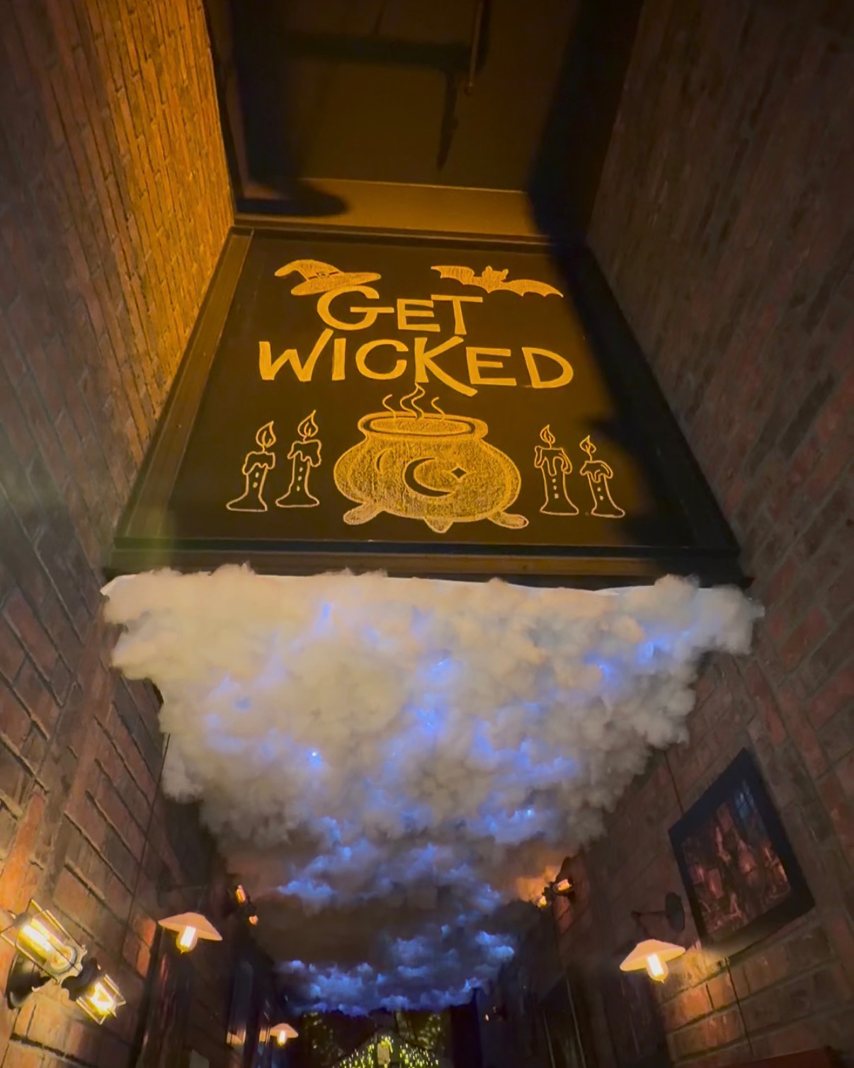 Get Wicked sign with a cauldron above a brick hallway filled with cloud-like Halloween ceiling decorations at Nason's.
