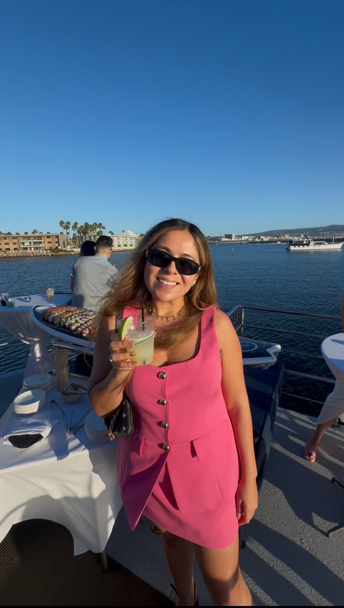 Giselle wearing a pink dress and sunglasses, holding a drink aboard a boat with the harbor behind her.