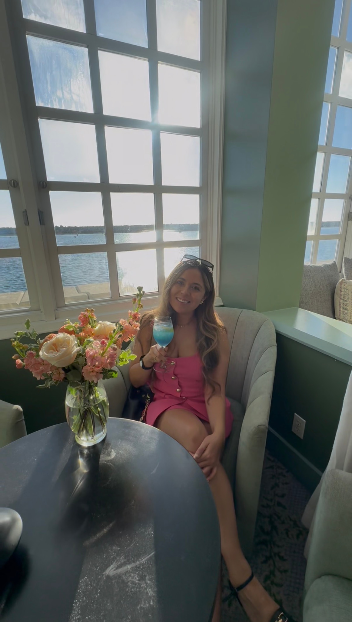 Giselle in a pink dress holding a blue cocktail beside a vase of flowers, with the harbor outside the lounge windows.