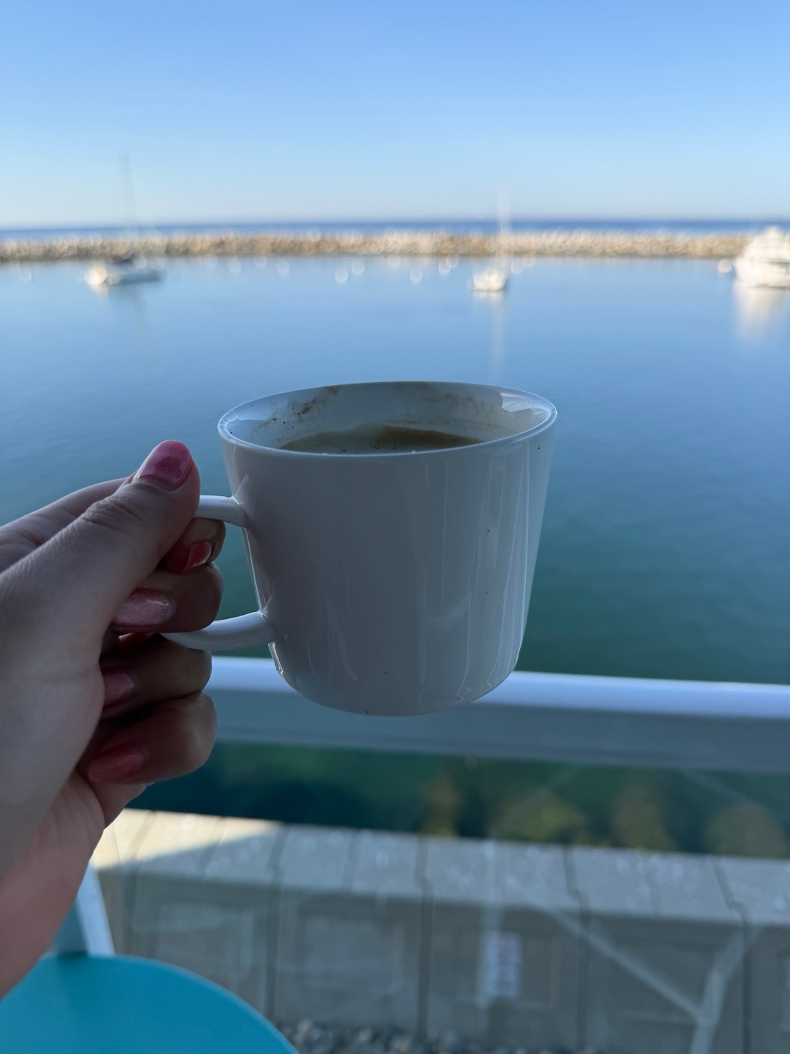 A hand holding a white coffee cup in front of a blue harbor with moored boats.
