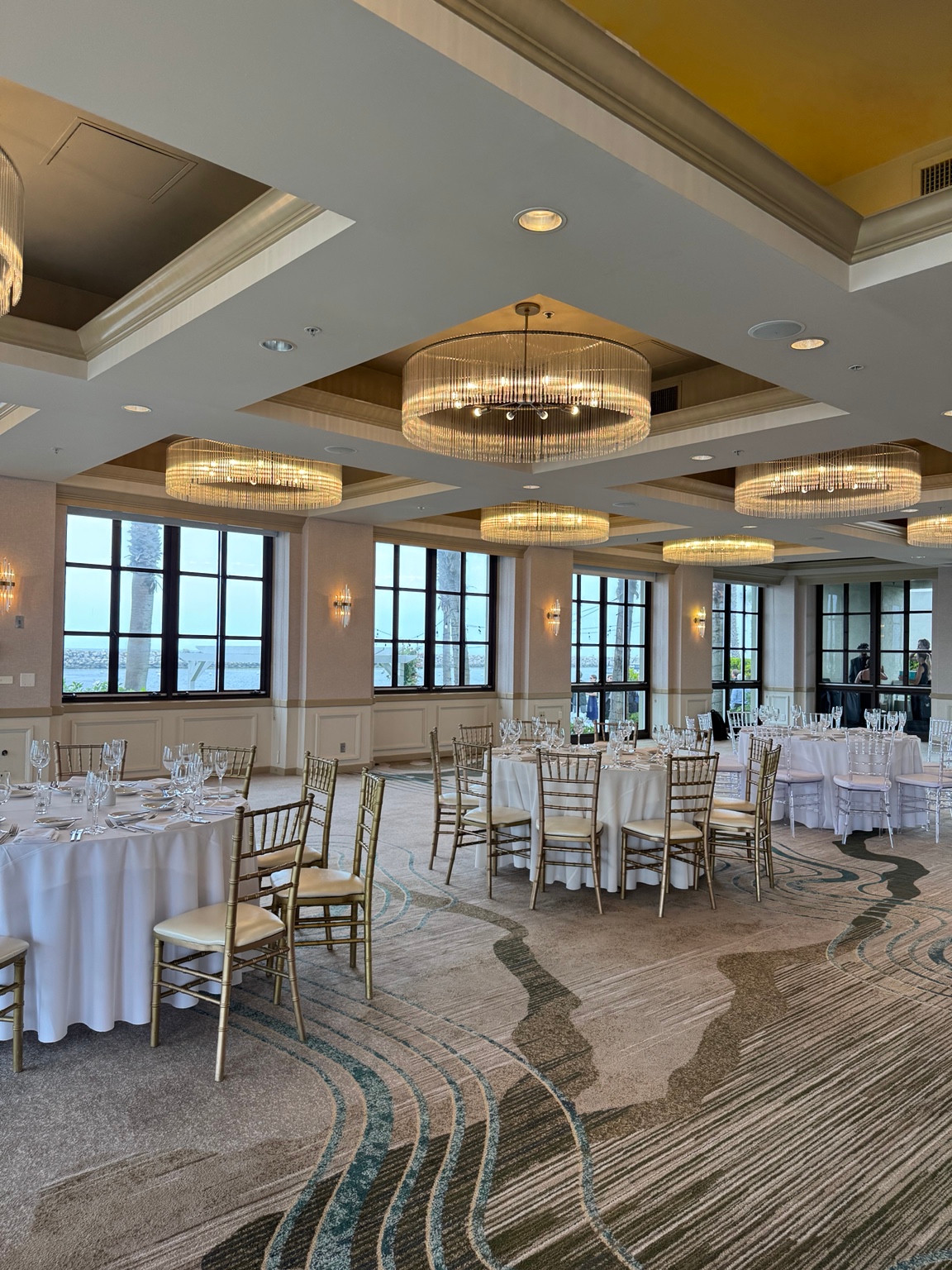 Round tables set with white linens and glassware beneath chandeliers in a ballroom with windows overlooking the water.