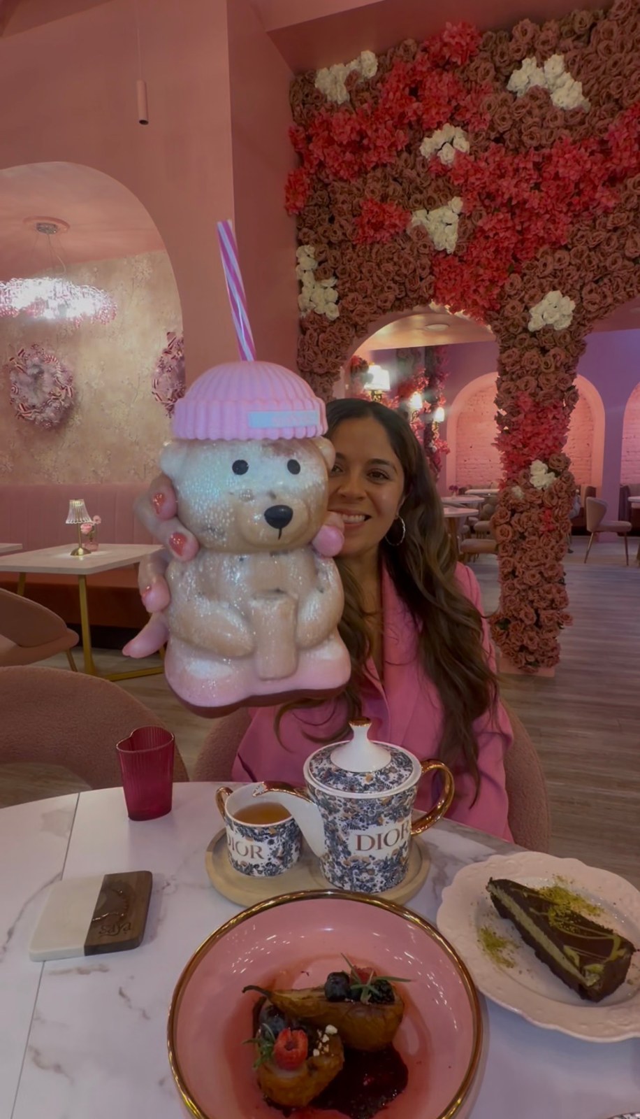 Giselle holding a teddy bear-shaped cup inside Saya Brasserie, surrounded by pink floral arches.