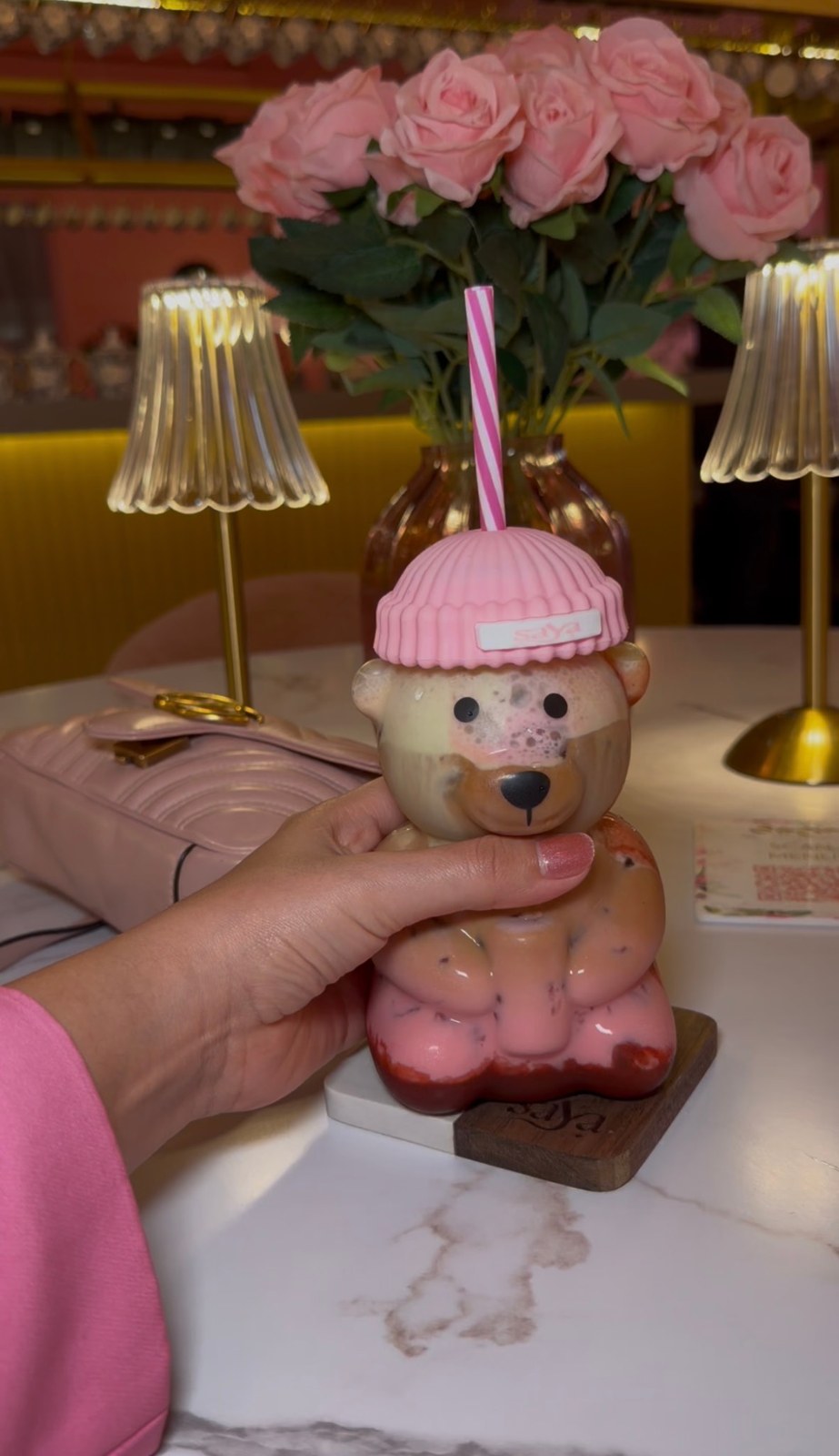 A hand holding Saya’s bear-shaped drink cup with a pink lid and striped straw, with pink roses behind it.