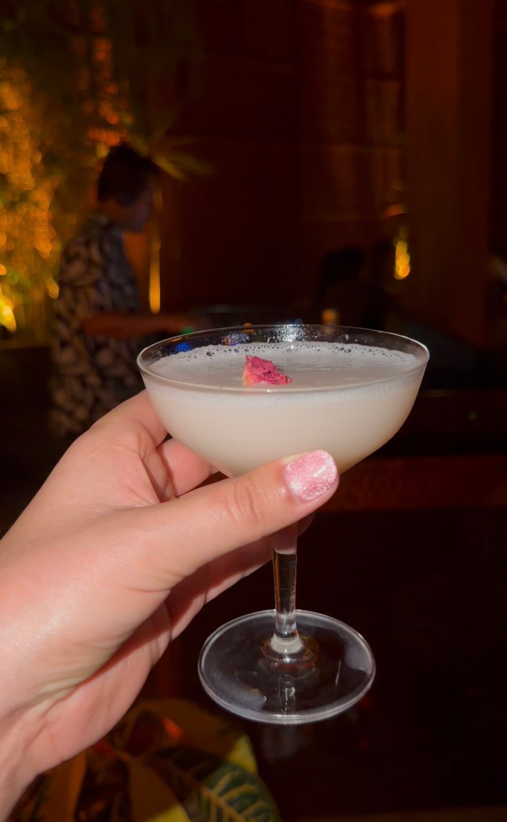 A pale cocktail in a coupe glass with a pink petal garnish against a warmly lit restaurant backdrop.