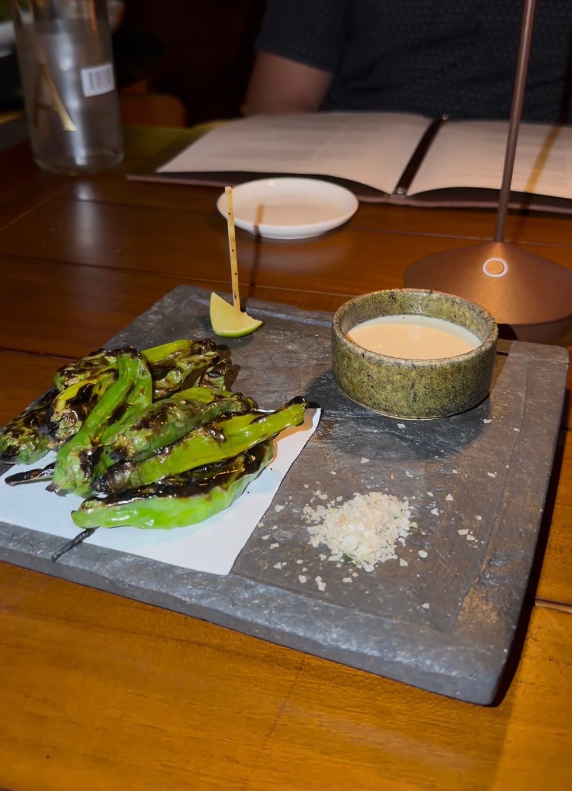 Charred shishito peppers on a slate platter with a lime wedge, seasoning and a bowl of dip.